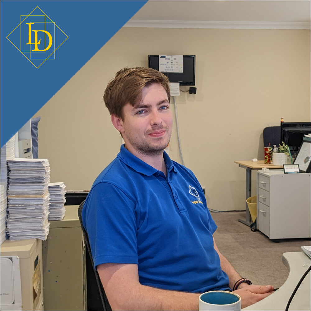 Aaron is a sitting at his desk with his hands in his lap smiling at the camera. Parts of the office can be seen in the background, some clutter is on his desk in the foreground. Up in the top left corner is the Logical Developments logo in gold on a blue background. The Logical Developments logo which is a diamond with the letters L and D inside of it. The letters are offset from each other vertically while also overlapping horizontally. The each sit inside of their own rectangle. 